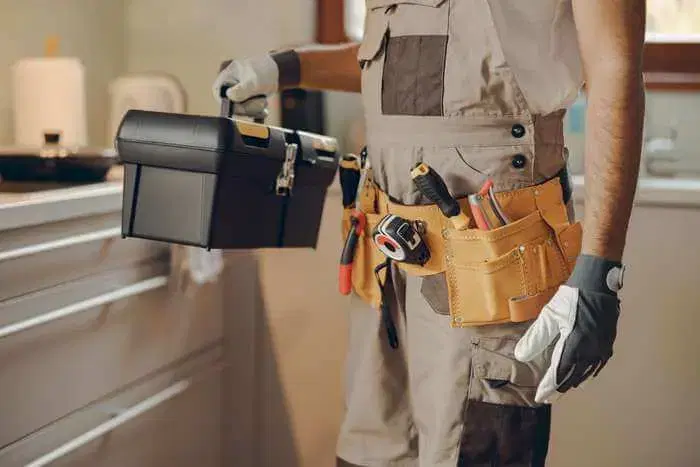 A tradesperson in overalls and a loaded tool belt carrying a toolbox through a kitchen.